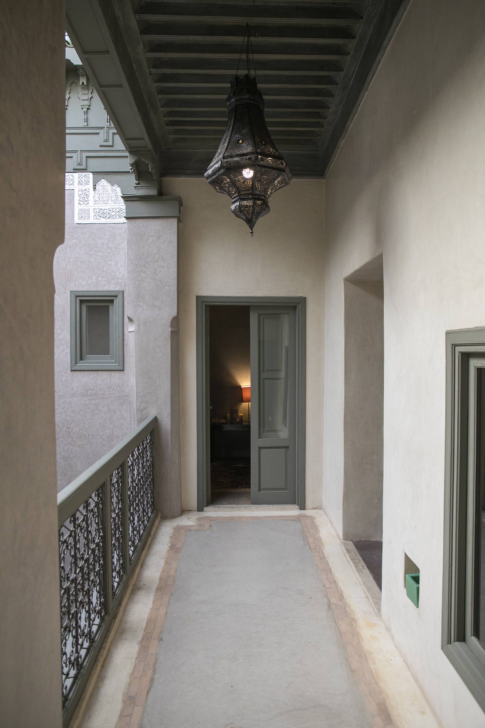 Au bout de la galerie du riad, la porte ouverte d'une chambre — une lampe allumée attend derrière, sous la lanterne de métal ajouré