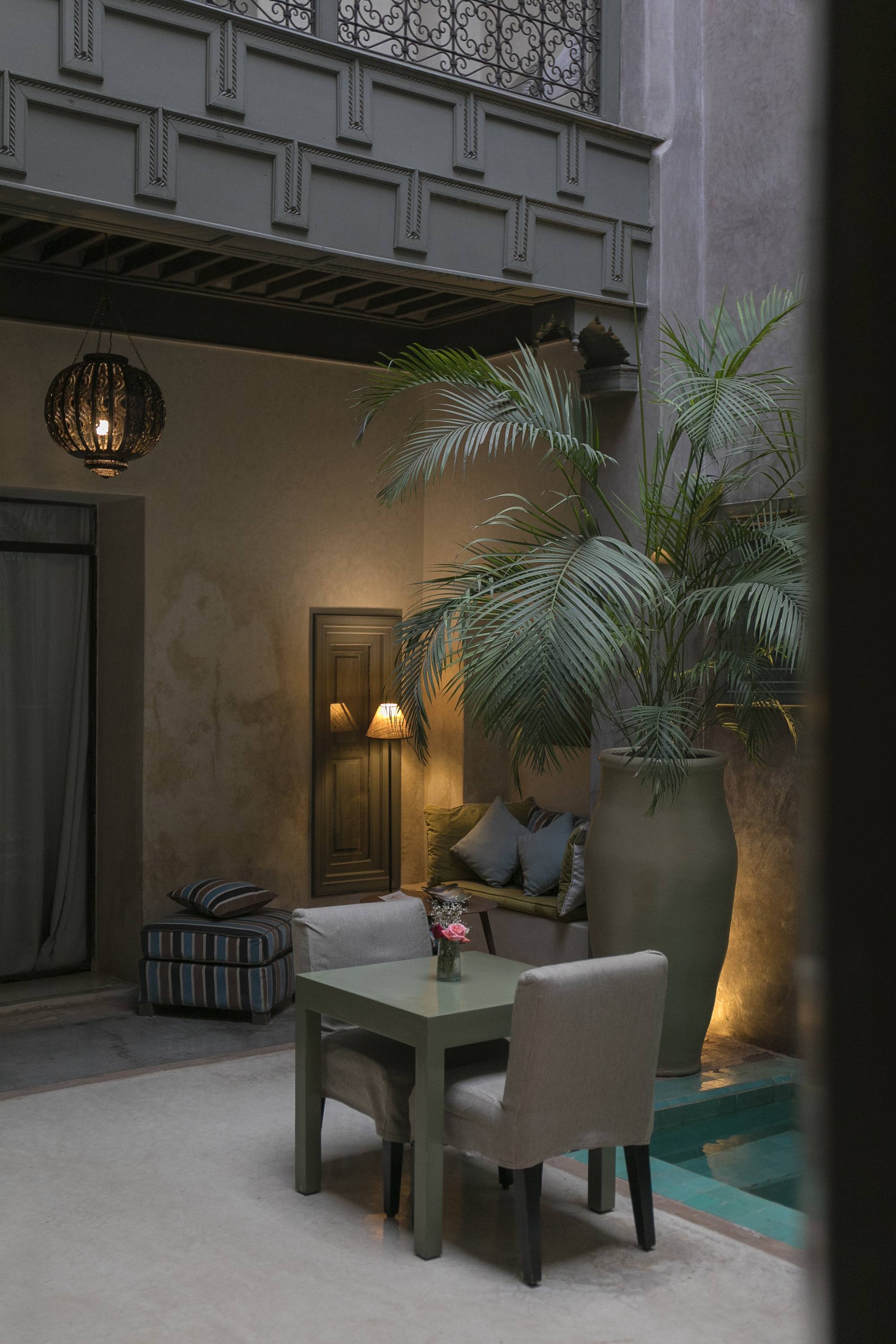 Un coin du patio le soir — la table pour deux sous la lanterne, le grand palmier et la piscine à ses pieds